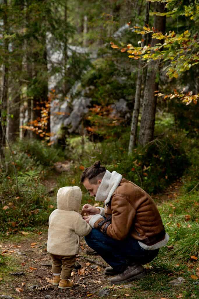 Father and toddle in a forest looking at something in the father's hands. 